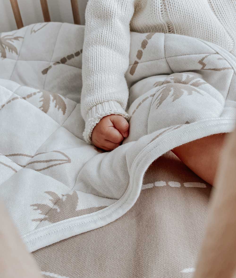 Baby's hand resting on a natural Surfing Palm reversible cotton quilt with sand palm and surfboard print
