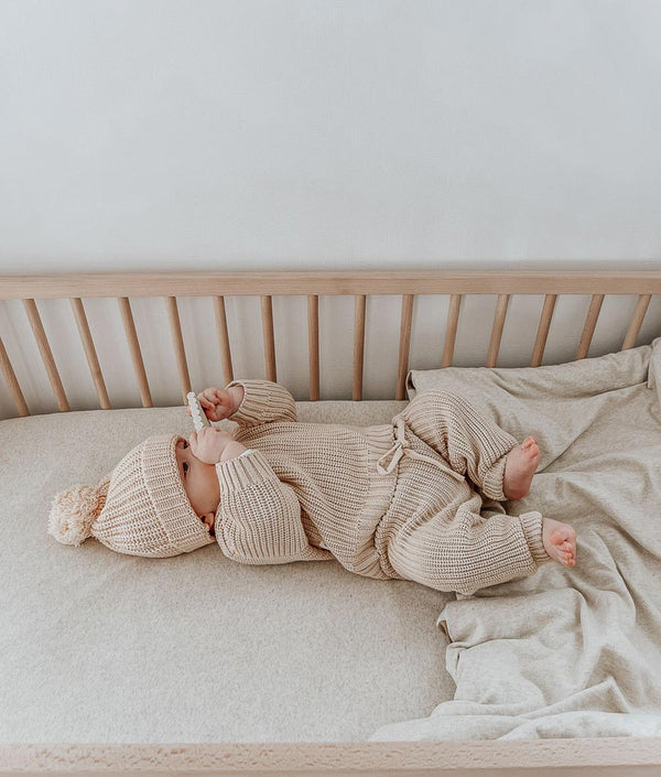 A baby laying in a crib with a JERSEY COT SHEET - NATURAL MARLE from Bengali Collections on.