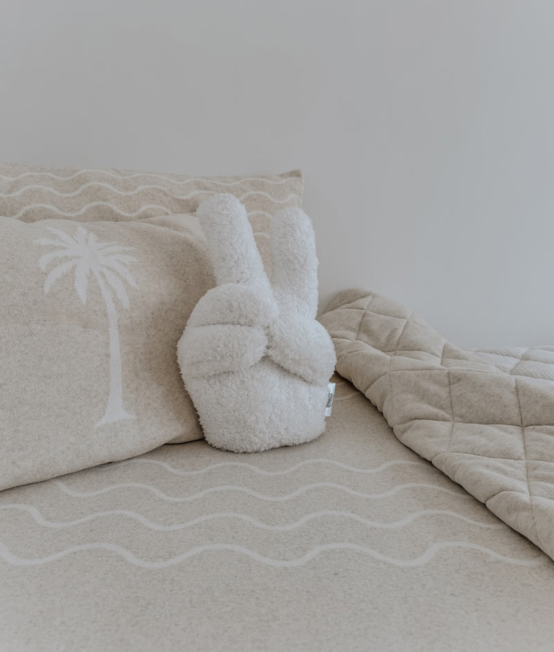Ivory Peace Sherpa cushion in a peace-sign hand shape on a bed with a palm tree pillowcase and wave sheet