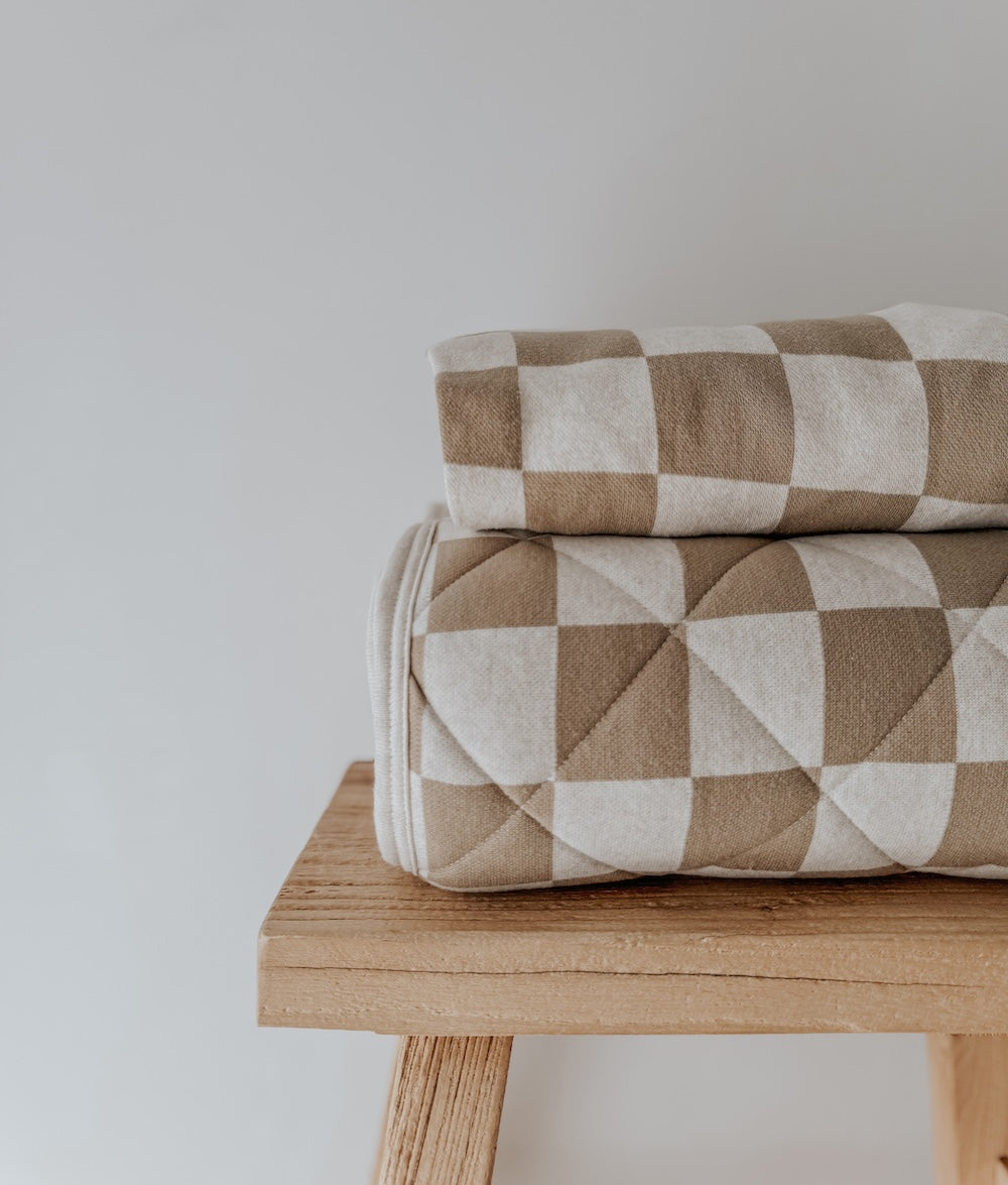 Khaki gingham quilt folded in two on a timber bench against a white wall