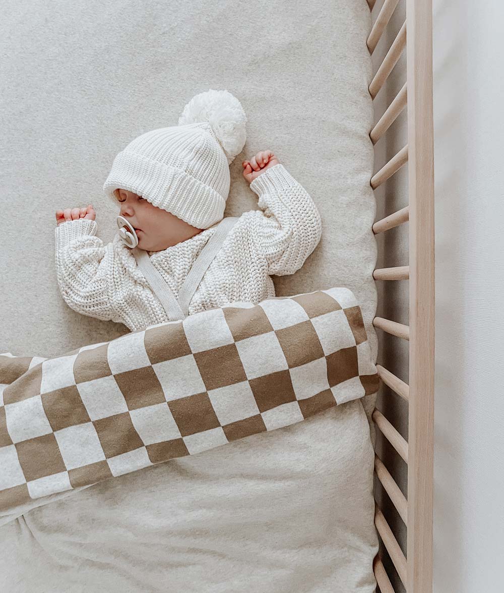 Baby in a knit beanie asleep in a timber cot under a khaki gingham jersey cotton duvet cover