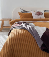 Bed with a tobacco quilt, palm print cushion and chocolate and ivory striped pillows behind