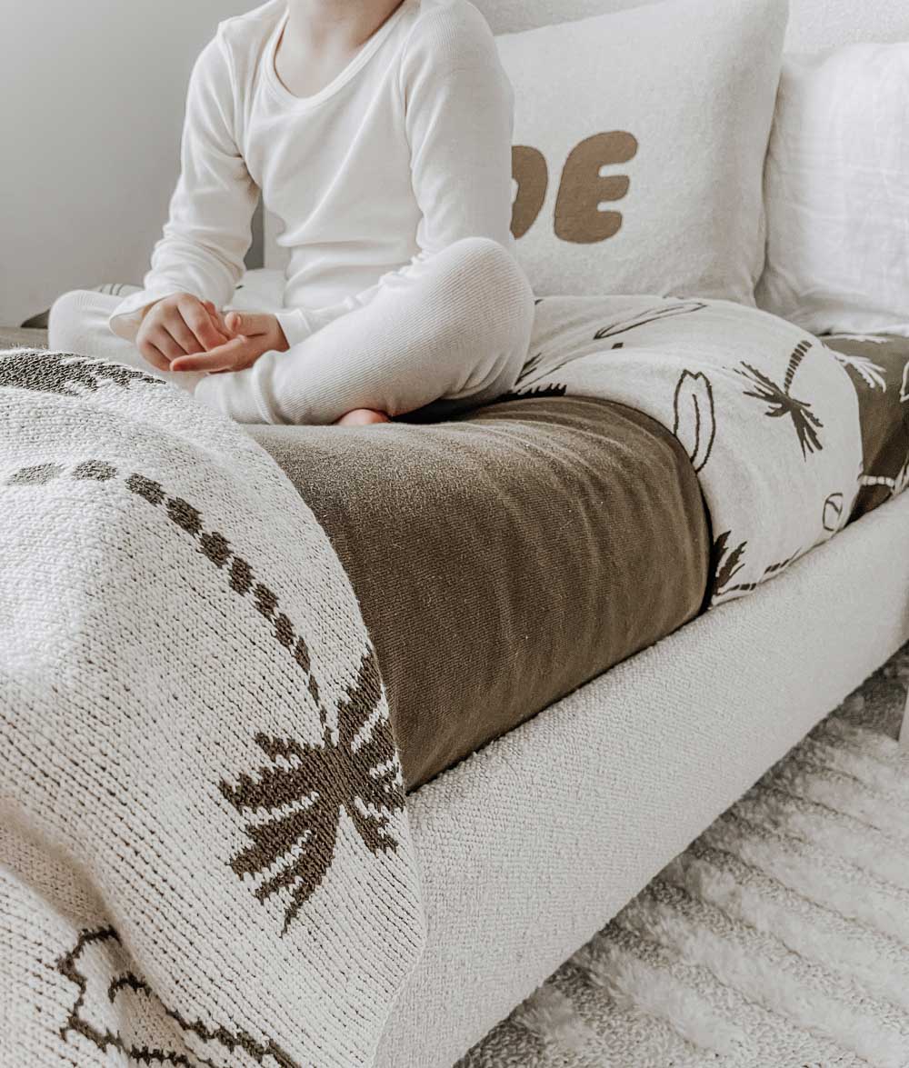 Child sitting cross-legged on a bed layered with a Surfing Palm duvet cover, olive sheet and palm knit blanket