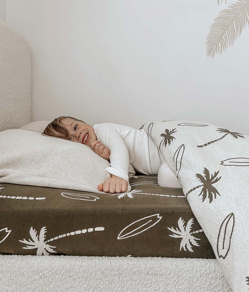 Smiling child resting on an olive Surfing Palm jersey fitted sheet with ivory palm and surfboard print