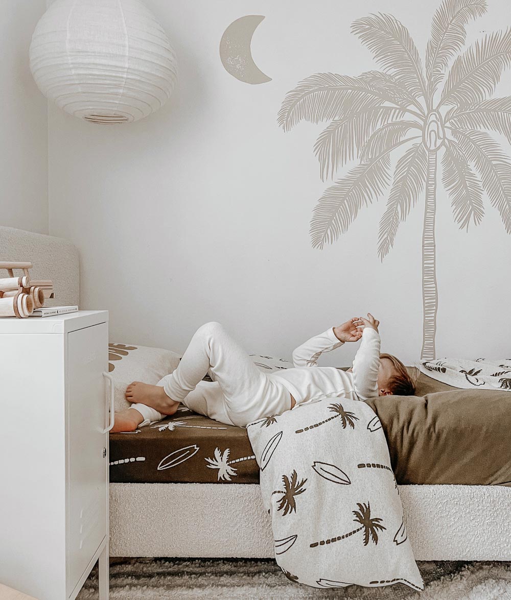 Child lying on a bed under a natural Surfing Palm kids duvet cover, olive fitted sheet and palm tree wall mural behind