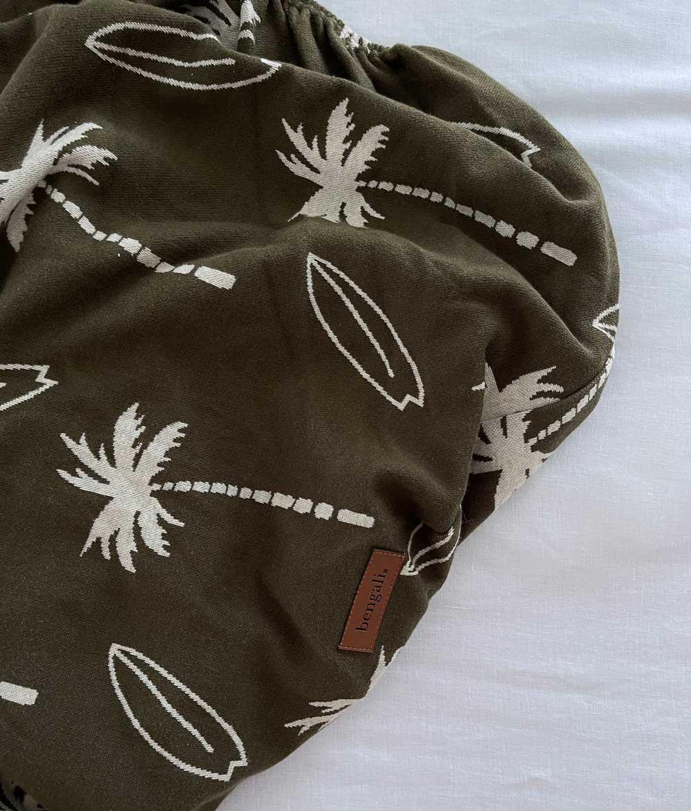 Olive Surfing Palm jersey fitted sheet gathered on white linen, showing the elasticised edge and leather label