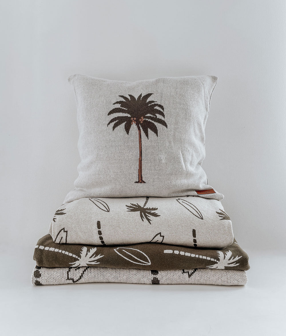 Stack of folded Surfing Palm kids bedding in natural and olive, topped with a palm tree cushion