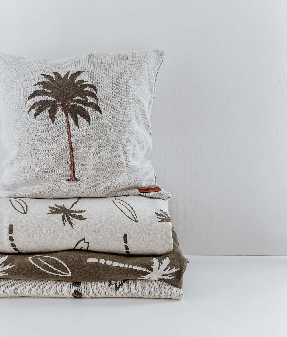 Stack of Surfing Palm knitted cotton baby blankets in natural and olive, topped with a palm tree cushion