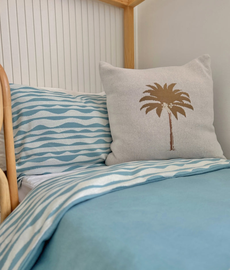 Khaki Palm cushion cover on a timber bed with a blue wave pillowcase and duvet cover