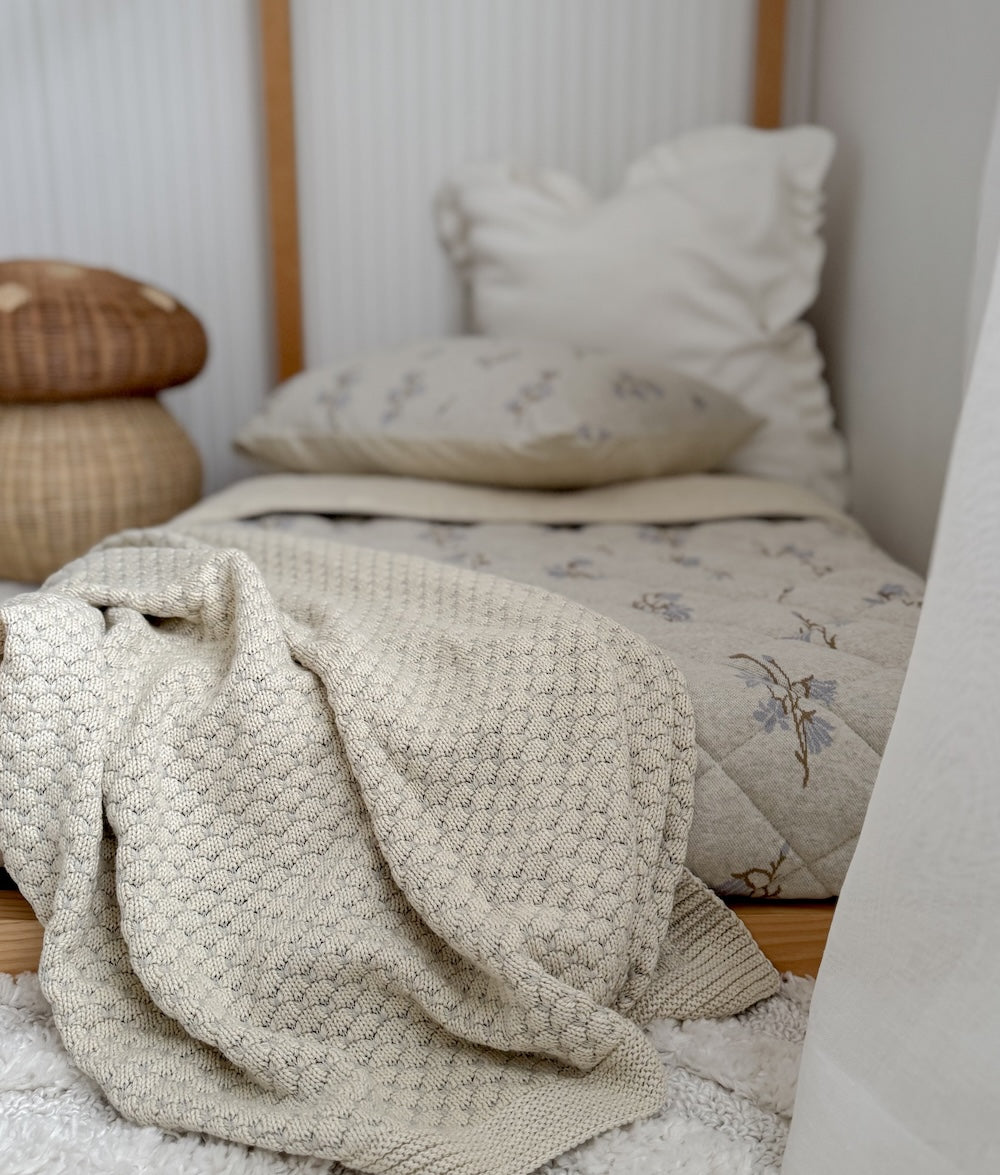 Pebble knit baby blanket in vanilla marle tumbled across a kids bed with floral bedding