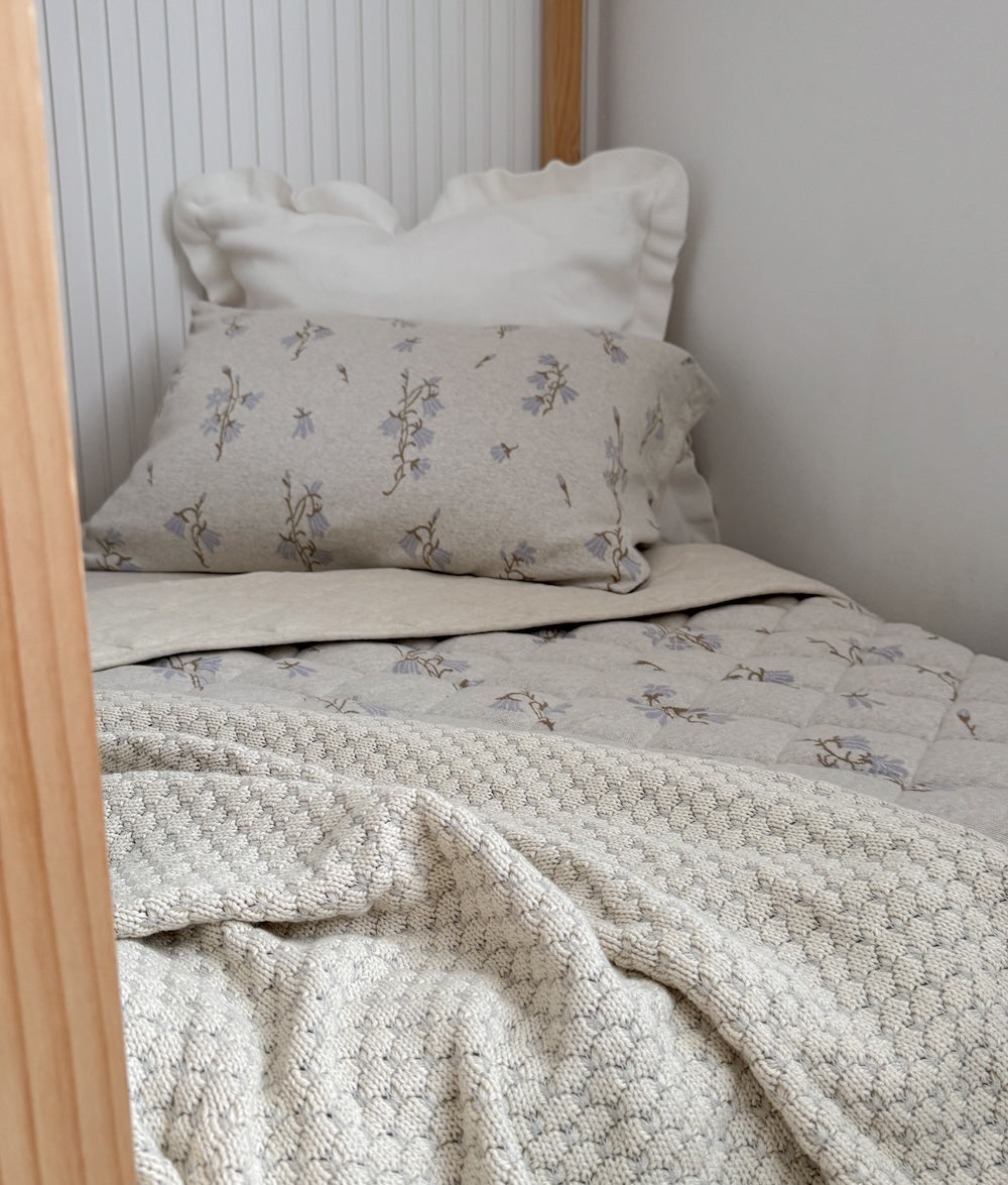 Vanilla marle knitted blanket over a toddler bed with a frilled cushion and floral pillowcase