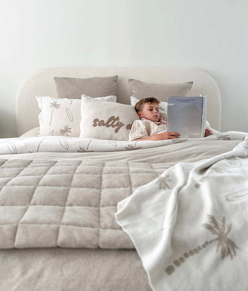Child reading in bed beside a Salty Days pillowcase, with palm print bedding and a greige quilt