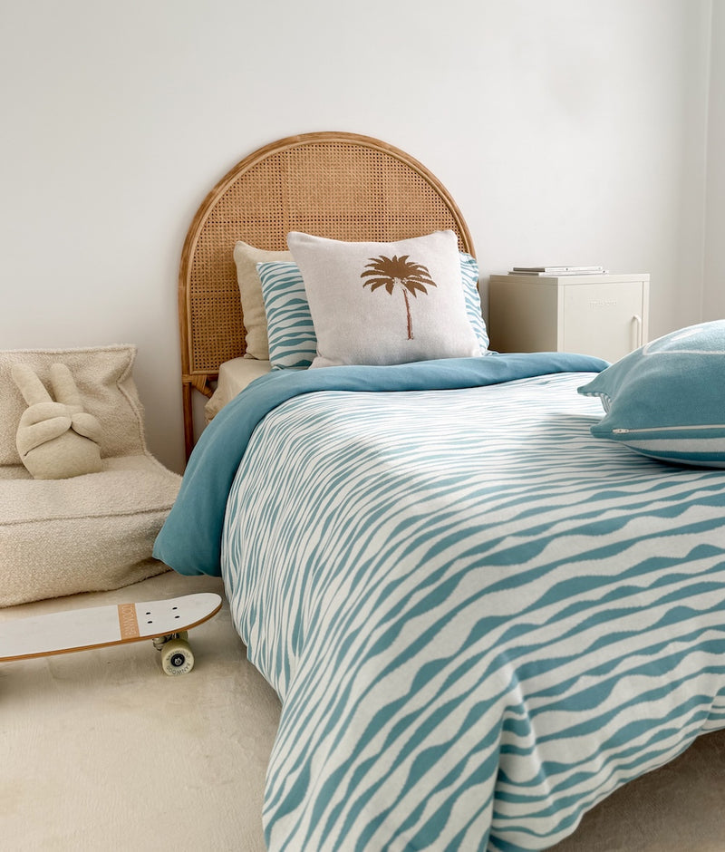 Kids bed with a rattan bedhead, blue wave bedding, the Khaki Palm cushion cover and a skateboard