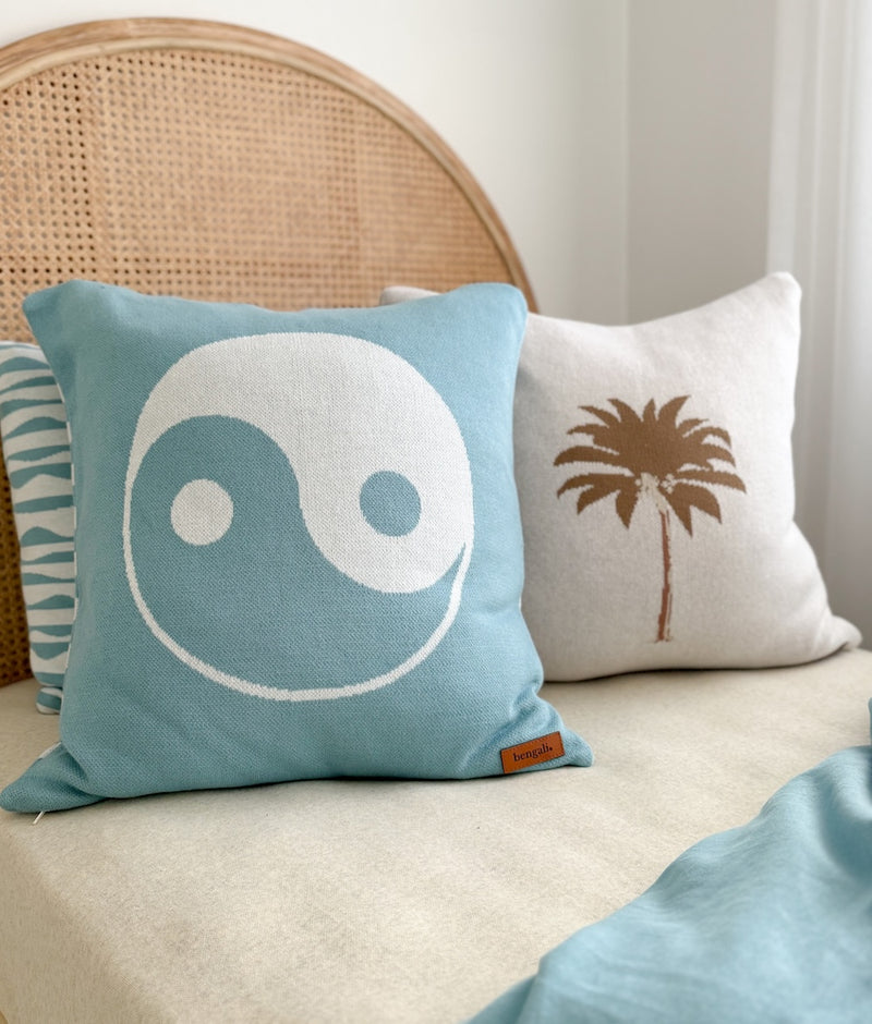 Khaki Palm cushion cover beside a blue yin yang cushion on a bed with a rattan bedhead