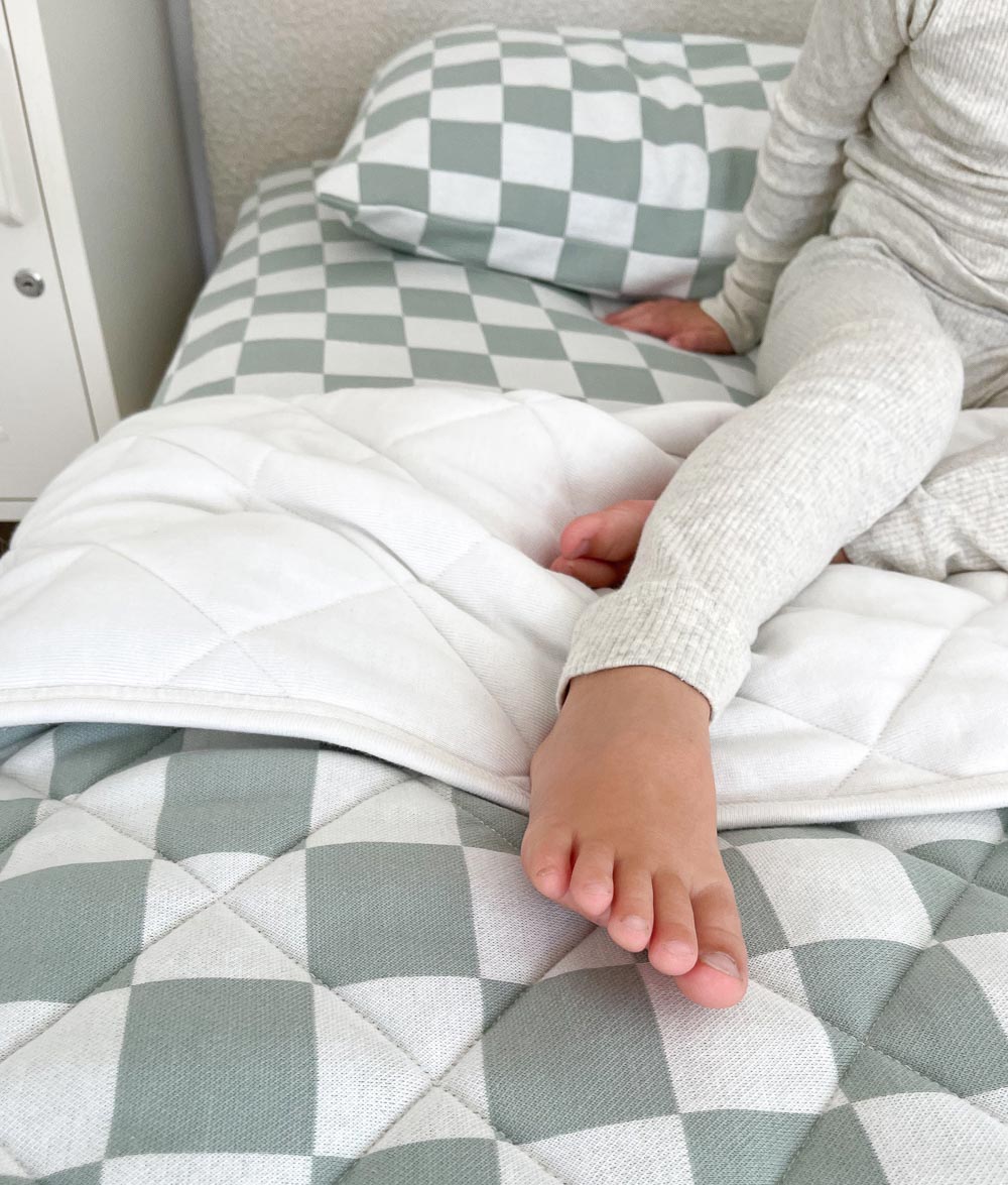 Child's leg resting on a Sage Gingham jersey fitted sheet beside a quilted sage gingham quilt
