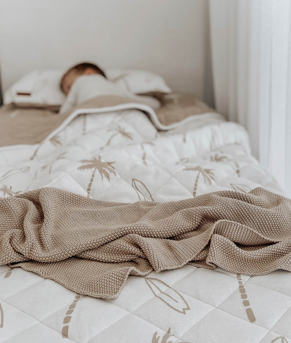 Child asleep under a natural Surfing Palm reversible kids quilt with a sand knit blanket across the foot