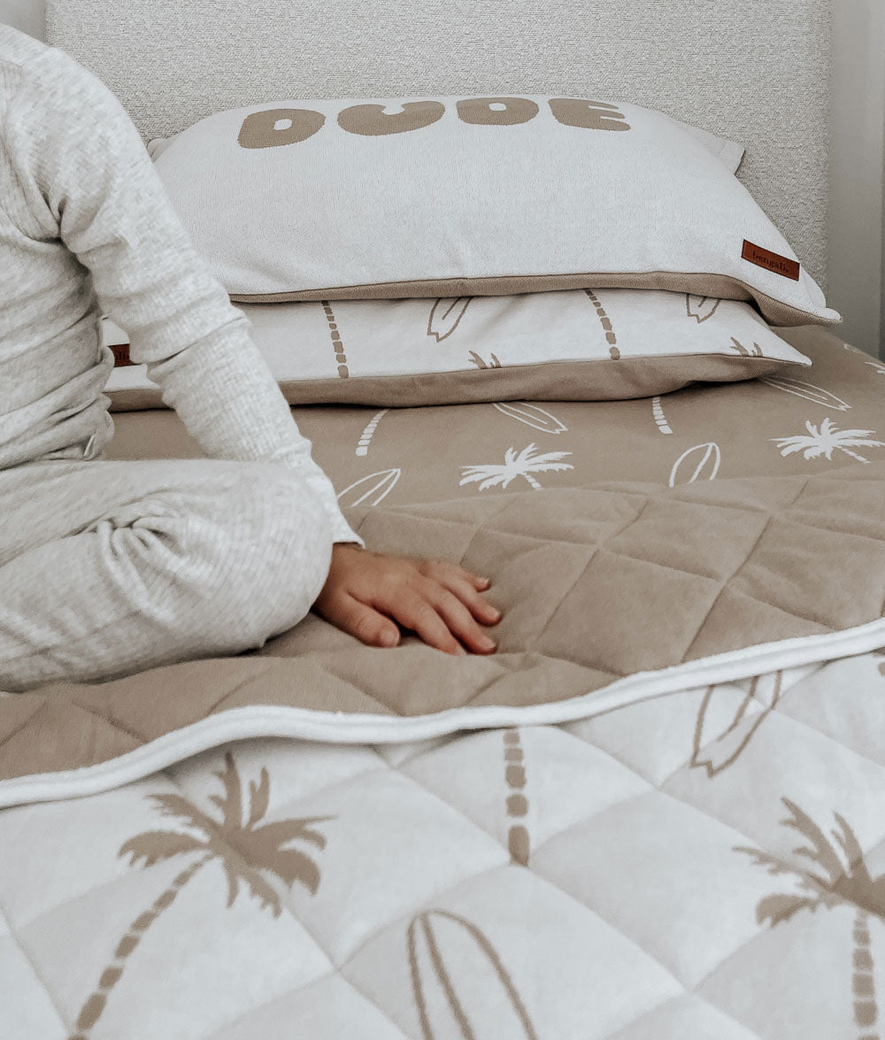 Child kneeling beside a DUDE jersey pillowcase on a bed with a quilted Surfing Palm quilt