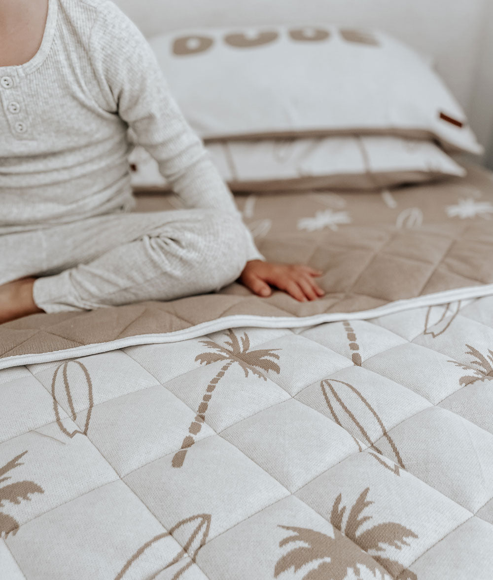 Child sitting on a bed with a Surfing Palm reversible quilt folded back to show its sand quilted side