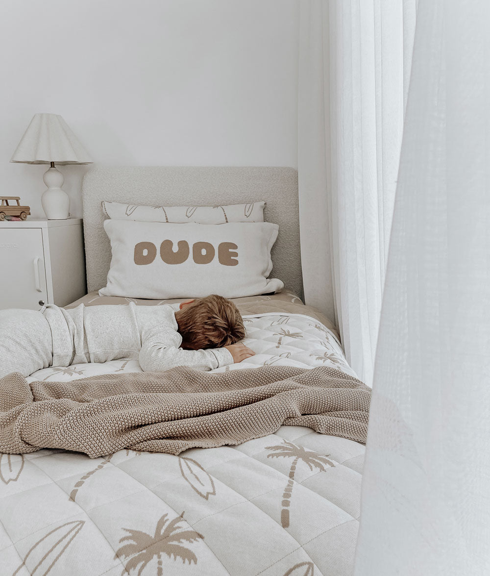 Child lying on a single bed below a DUDE jersey pillowcase with sand lettering, over a Surfing Palm quilt