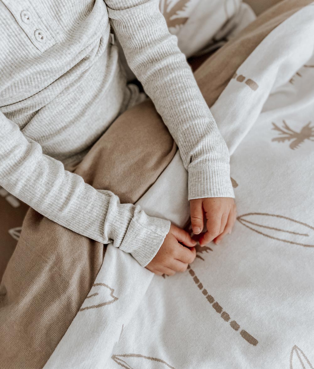 Child in a knit top sitting on a Surfing Palm kids duvet cover in the natural colourway with sand palm print