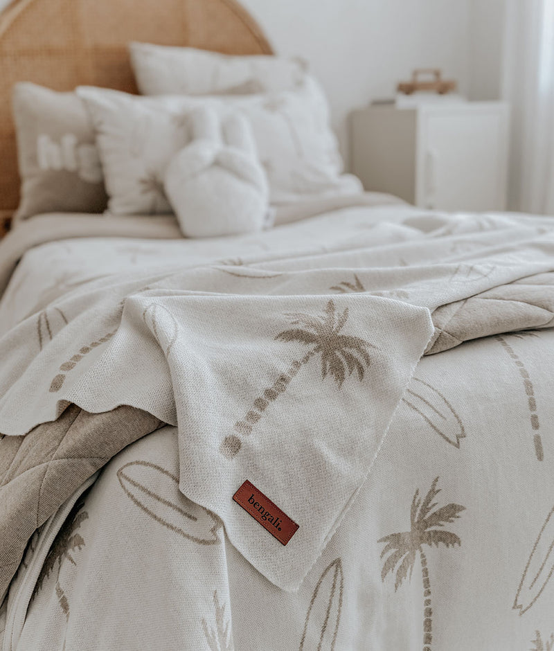 Surfing Palm knitted cotton baby blanket in dune draped over a kids bed with a rattan bedhead and cushions