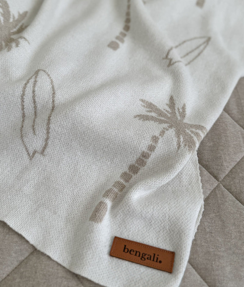 Corner of a dune Surfing Palm knitted cotton baby blanket with leather label on a greige quilted throw