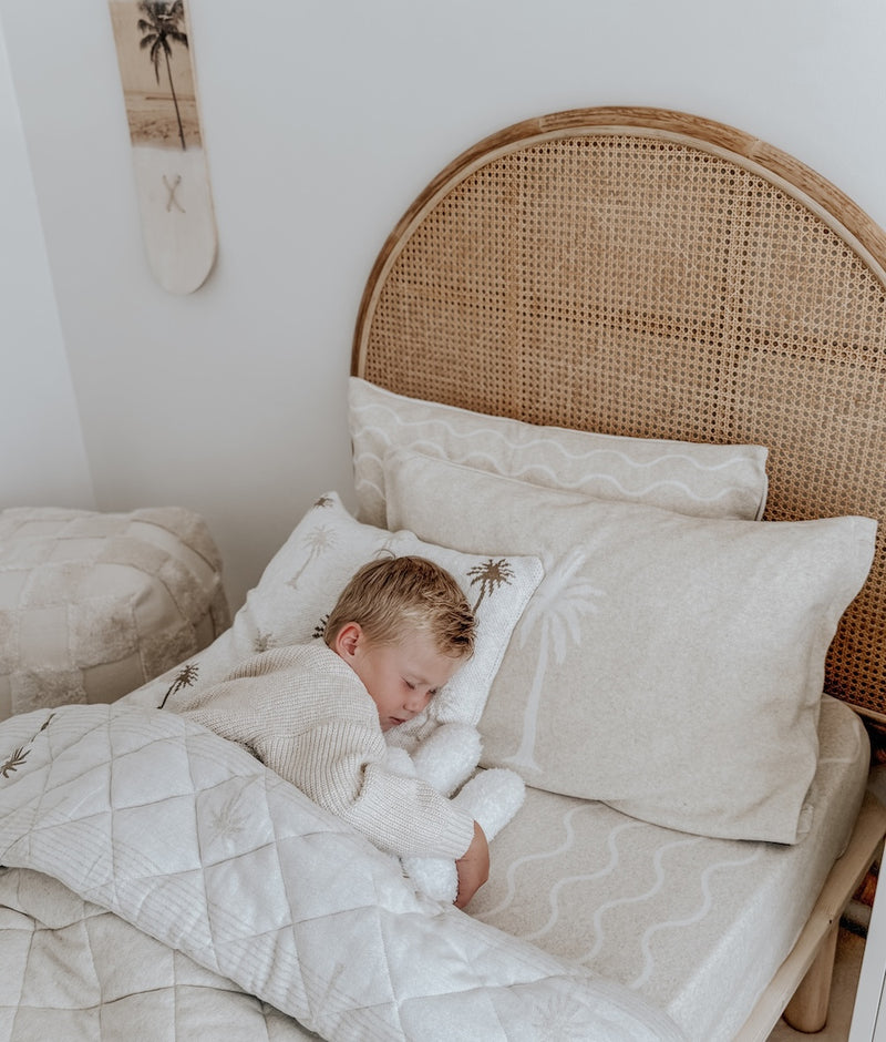 Child asleep on a Coastal Marle jersey fitted sheet under a quilt, with a rattan bedhead behind