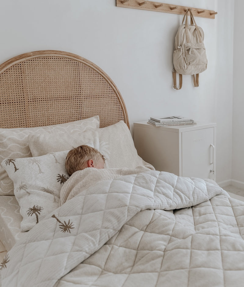 Child asleep on a kids bed with the Palma palm cushion, quilted bedding and a rattan bedhead