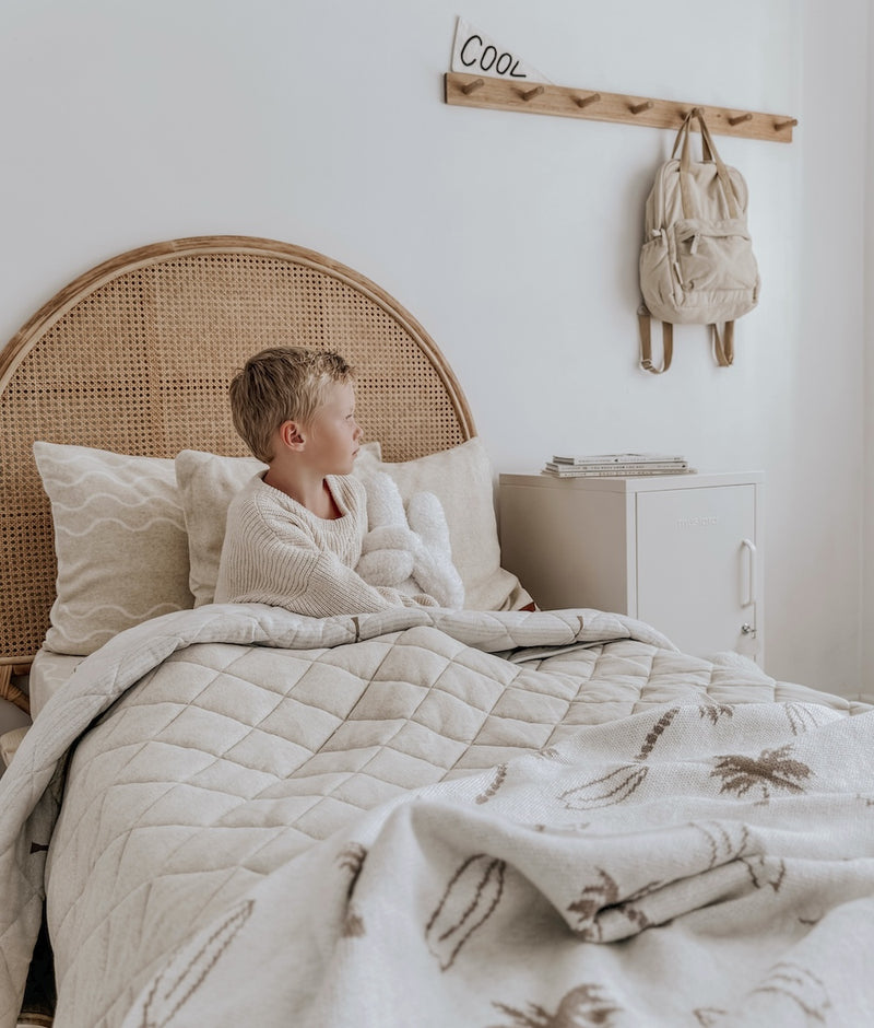 Child sitting up in a kids bed with ivory pillowcases, a palm print quilt and a rattan bedhead
