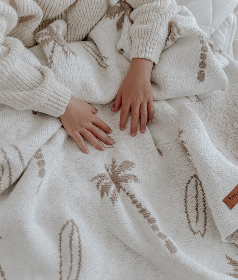 Child's hands resting on a dune Surfing Palm knitted cotton baby blanket with sand palm and surfboard print