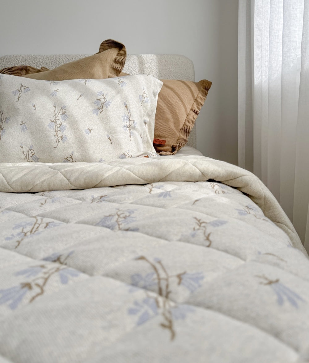 Bed with floral quilted bedding and khaki frill euro pillowcases beside a sheer curtain