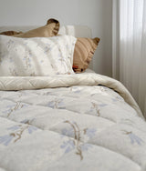 Bed with floral quilted bedding and khaki frill euro pillowcases beside a sheer curtain