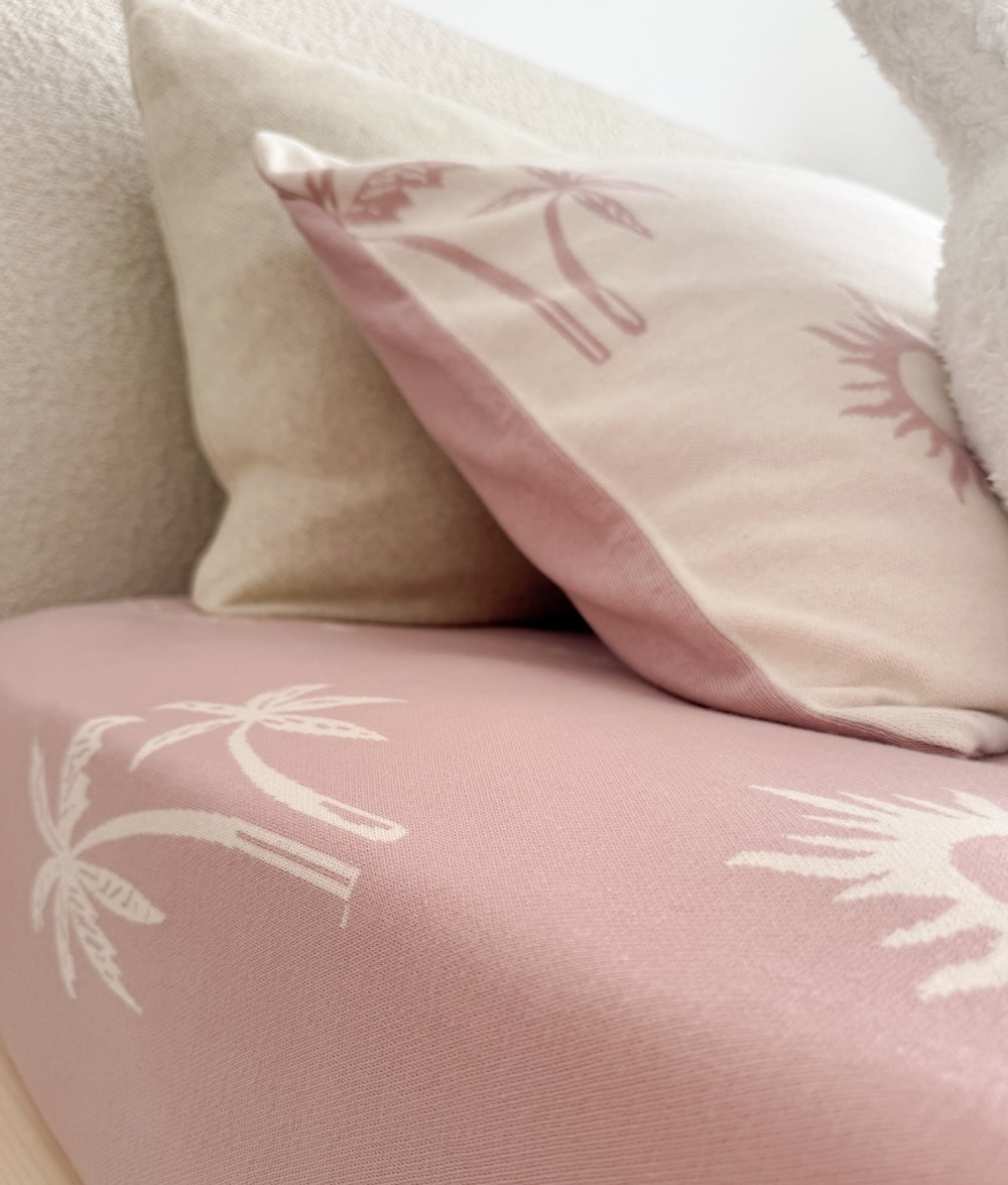 Cream palm and sun print pillow resting on a soft pink fitted sheet