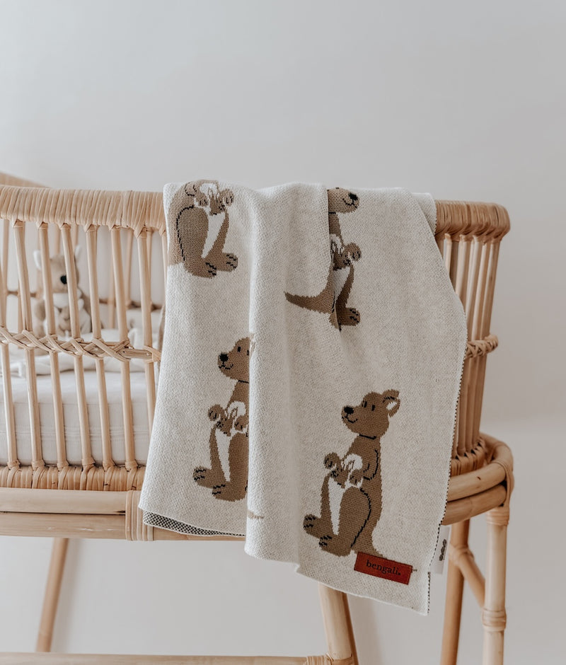 Kangaroo knitted cotton baby blanket draped over a rattan bassinet, showing khaki kangaroos and joeys