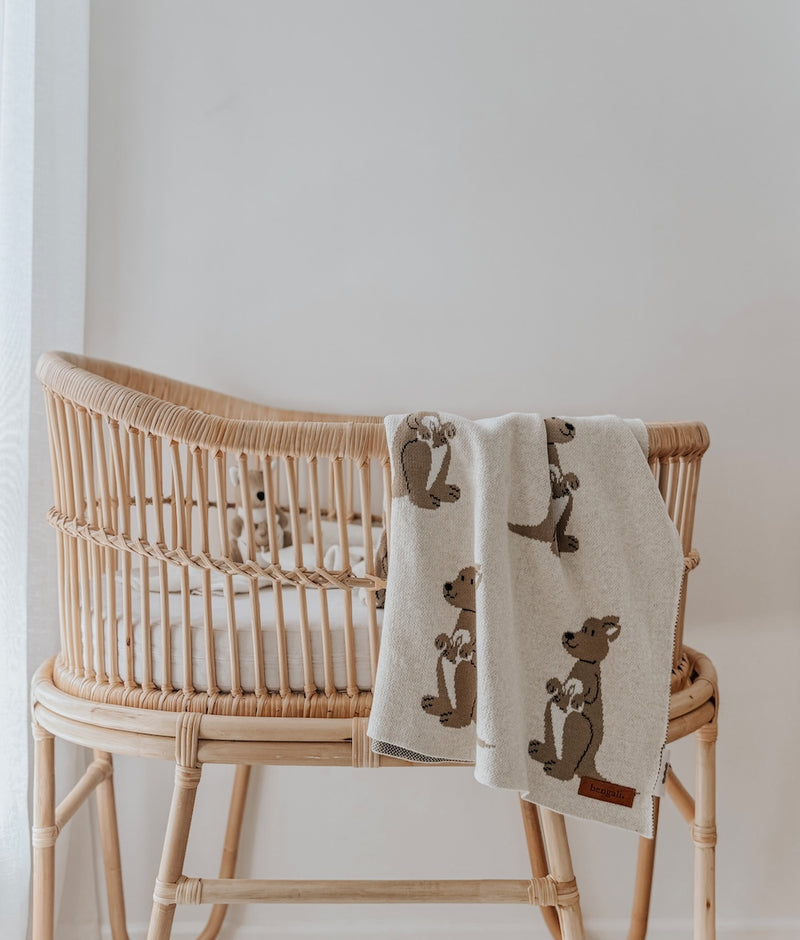 Kangaroo knitted cotton baby blanket hung over the side of a rattan bassinet on a stand