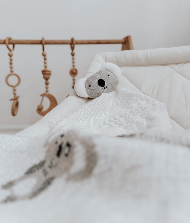 Koala baby comforter snuggly tucked into a bassinet beside a timber play gym