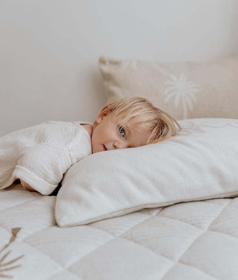 Child lying on an ivory cotton pillowcase with a palm tree pillowcase behind, on a quilted bed