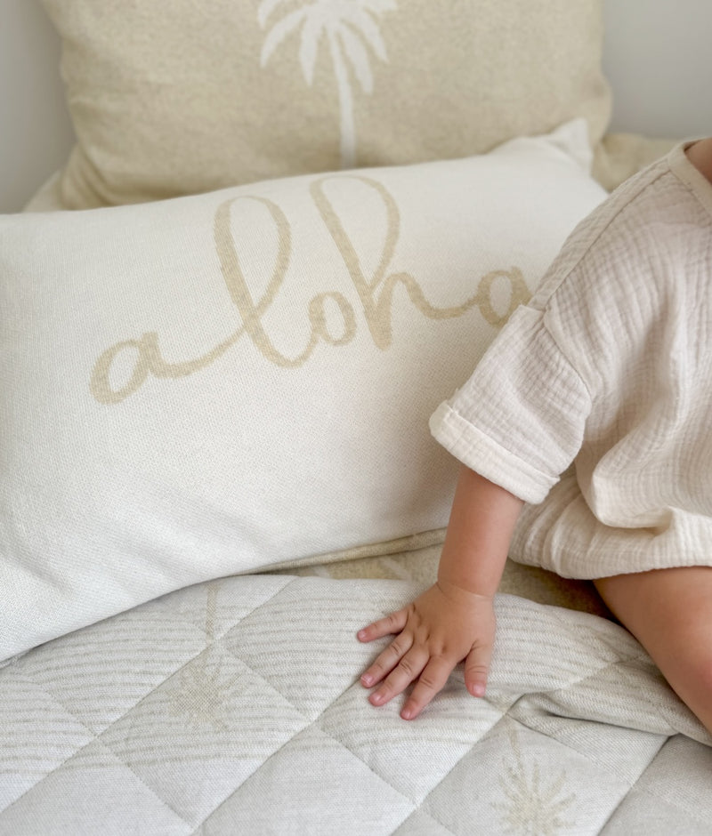 Baby's hand on a quilted bed beside the Aloha cotton pillowcase with natural script lettering
