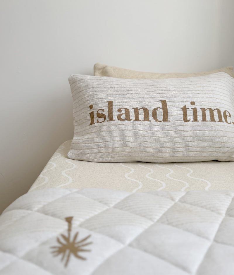 Close view of the Island Time pillowcase's khaki lettering on a bed with a wave print sheet and palm quilt
