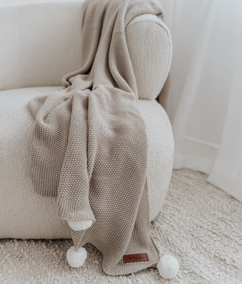 Greige Pom-Pom knit cotton throw with ivory pom poms draped over a curved boucle armchair