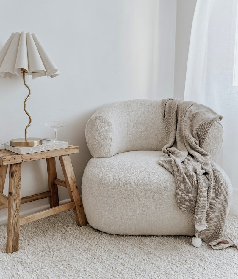 Greige Pom-Pom lounge throw draped over a boucle armchair beside a timber side table and pleated lamp
