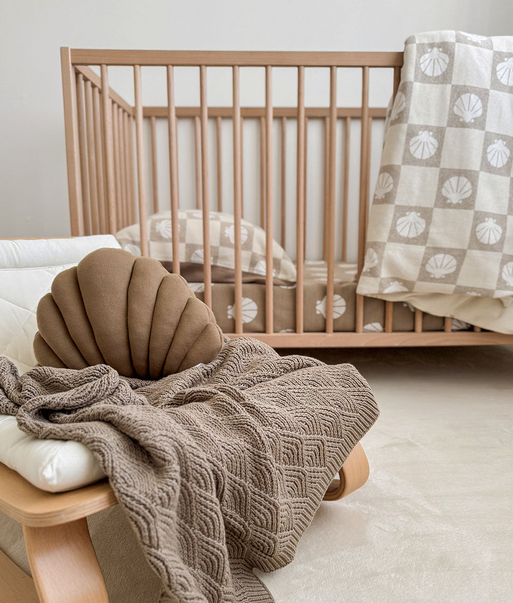 Stone shell stitch blanket draped over a timber bench beside a cot with a shell cushion