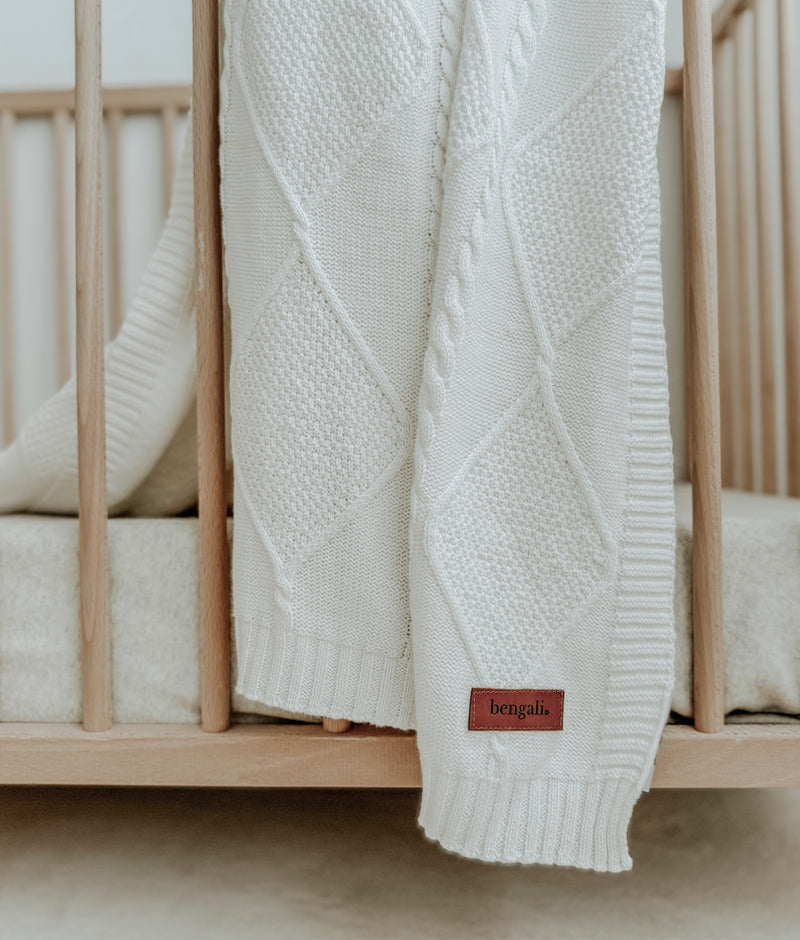 Close-up of the Ivory Diamante lounge throw's diamond and cable knit over a cot rail, with its leather label
