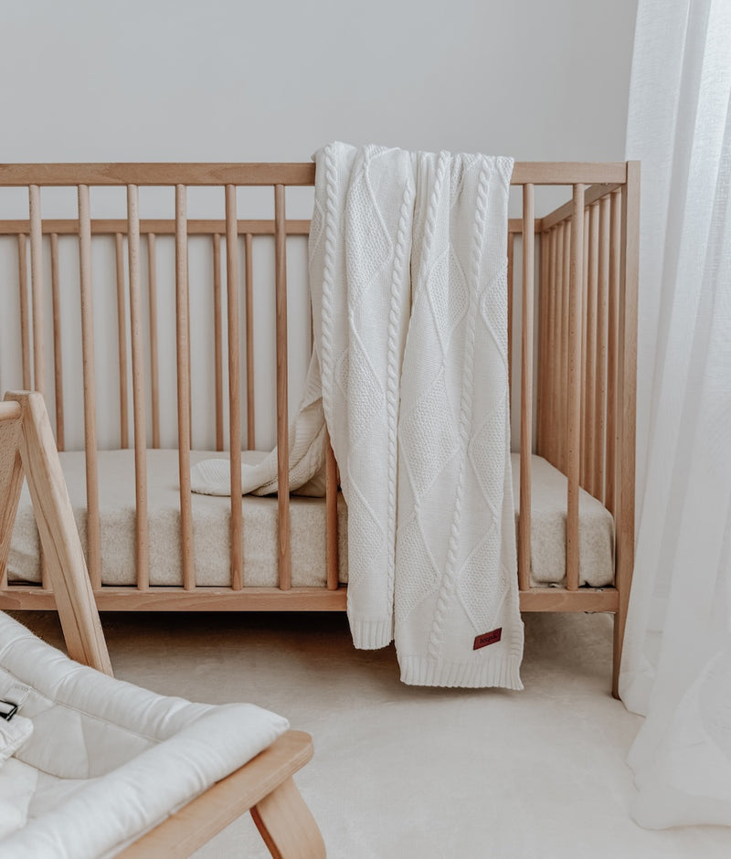 Ivory Diamante knit cotton throw over a timber cot rail, a baby bouncer in the foreground