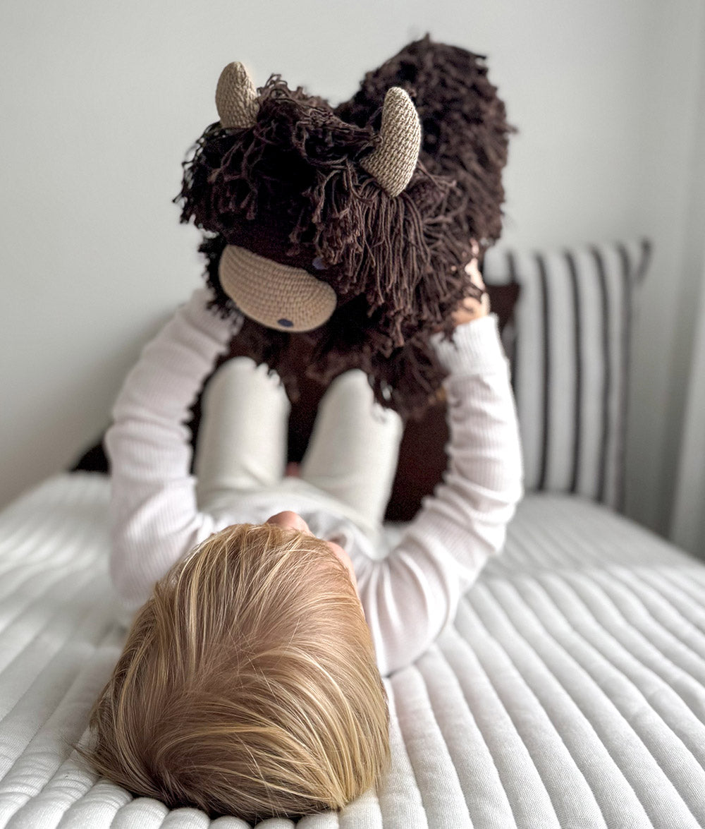 Child lying on a ribbed ivory quilt holding up Fergus the chocolate Highland cow soft toy