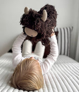 Child lying on a ribbed ivory quilt holding up Fergus the chocolate Highland cow soft toy