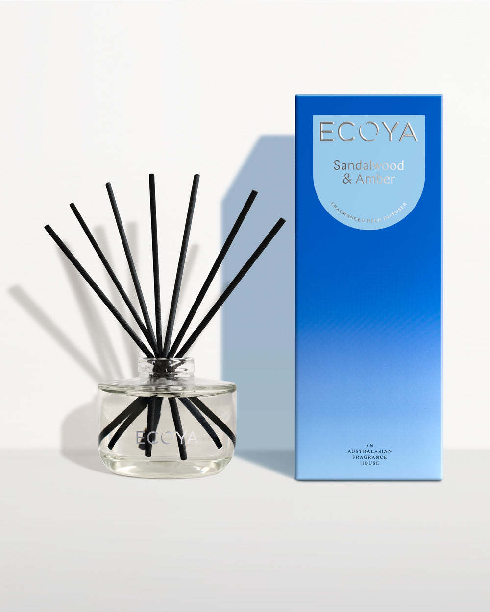 Ecoya Sandalwood and Amber reed diffuser in a clear glass jar with black reeds, beside its blue gift box