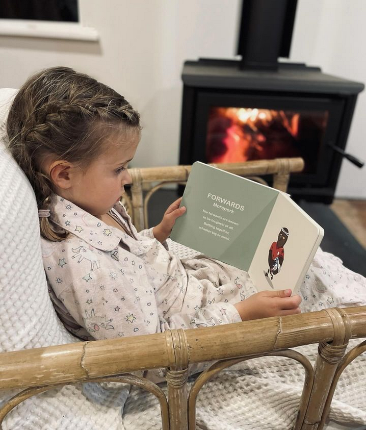 Girl reading Wild About Footy in a cane chair beside a lit wood burner