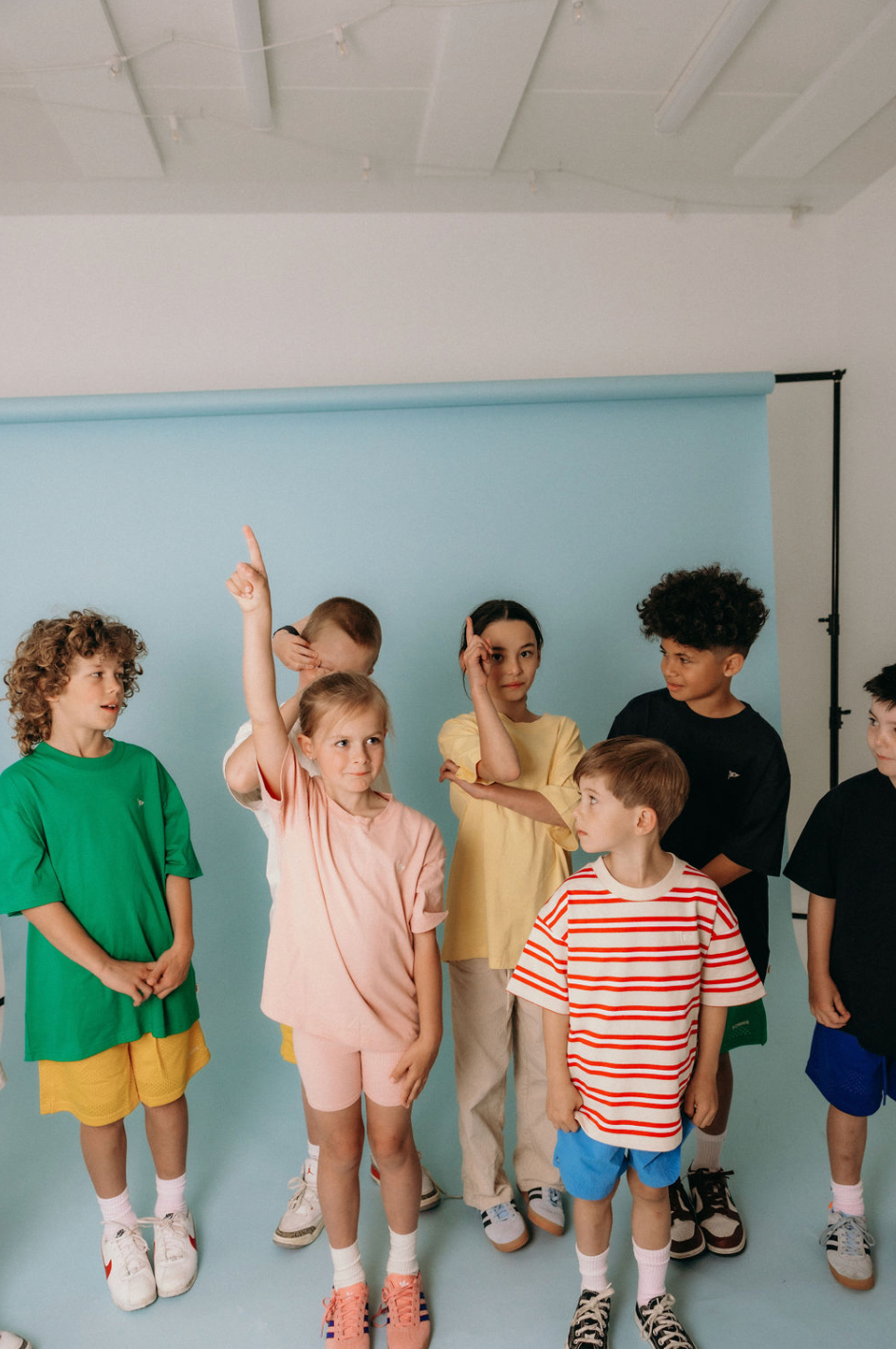 A group of children standing against a pale blue backdrop, one with a hand raised