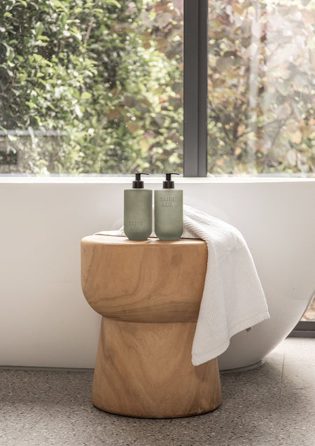 Two sage Smith & Co. bottles on a timber stool beside a freestanding bath and white towel